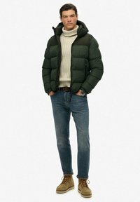 Grøn dunjakke med hætte, cremefarvet turtleneck, blå jeans og lysebrune snørebåndsstøvler. Har en puffy tekstur og en kontrastfarvet skulderpanel.