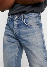 Džínové kalhoty ve vybledlé modré barvě se rovný střihem, viditelným šitím, pěti kapsami a stříbrným zapínáním na knoflík.