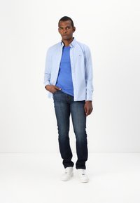 Camisa de cuadros azul claro sobre una camiseta azul, combinada con jeans oscuros y zapatillas blancas. Look simple y casual.