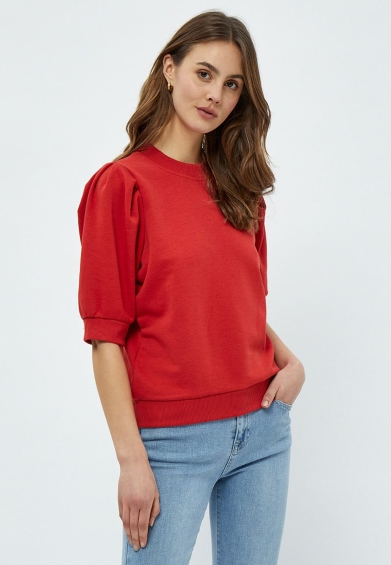 Röd sweatshirt med puffärmar, rund halsringning och ribbad nederkant. Bärs över ljusblå jeans med en slät textur.