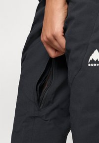 Pantalonii de zăpadă negri au o buzunar cu fermoar cu interior din plasă, o textură de țesătură netedă și un logo vizibil cu grafică montană pe lateral.