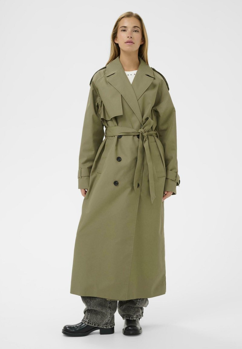Femme portant un long trench-coat olive ceinturé par-dessus un jean gris ample et des bottes noires, se tenant contre un fond blanc uni.