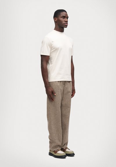 Bărbat în picioare, îmbrăcat cu o cămașă simplă de culoare albă-crem cu mânecă scurtă, pantaloni texturati maro și mocasini verzi și maro cu accente de lanț aurit.