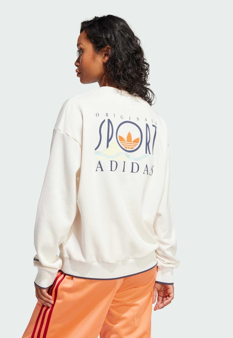 Bijela dukserica s grafikom "SPORT" i "ADIDAS" na leđima, s rebrastim manžetama. Uparena s narančastim satenskim hlačama i vidljivim detaljem logotipa.