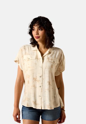 Frau mit dunkel-lockigem Haar, die ein beige-farbenes, kurzärmeliges Hemd mit Westernmuster und Jeansshorts trägt und zur Seite blickt.