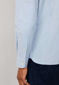 Camicia a quadri blu chiaro con toppe sui gomiti, polsini con bottone e orlo curvo. Il tessuto ha una consistenza liscia e presenta dettagli di pattern sottili.