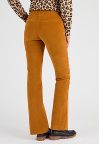Pantalons en velours côtelé évasés de couleur jaune moutarde, présentant une texture douce, des poches arrière et une apparence lisse, associés à des chaussures noires.