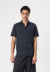Chemise à manches courtes, bleu marine foncé, à boutons, avec un motif texturé, une poche poitrine et des boutons marron, associée à un pantalon large foncé.