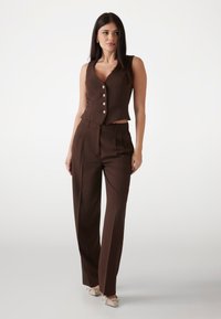 Bruine op maat gemaakte vest met parelknoopjes en een cropped ontwerp, gecombineerd met bruin, hoog-waist broeken. Kenmerkt zich door een soepele stof en strakke lijnen.