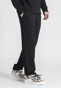 Zwarte gebreide broek met een gestructureerd kabelpatroon, voorzien van zijzakken en een relaxte fit, gecombineerd met gedessineerde sneakers.