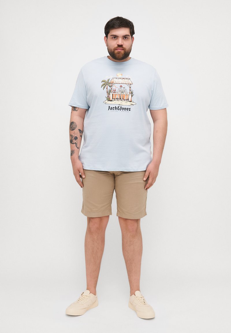Hellblaues T-Shirt mit kurzen Ärmeln und einer Tropengetränk-Illustration, kombiniert mit beigen Shorts. Weiße Turnschuhe runden das Outfit ab.