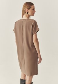 Braunfarbenes Kurzarmkleid mit gerader Silhouette, glatter Textur und einer Naht in der Mitte des Rückens. Kombiniert mit kniehohen schwarzen Stiefeln.