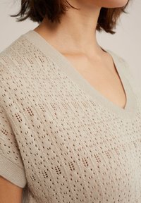 Ljusbeige stickad tröja med v-ringning, detaljerat hålmönster och ribbade kanter som framhäver en texturerad yta.