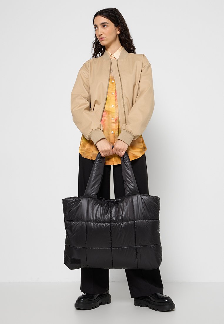 Frau mit langen dunklen Haaren trägt eine beige Jacke und ein gemustertes Hemd und hält eine große schwarze gesteppten Tragetasche vor einem weißen Hintergrund.