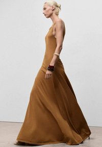 Largo vestido marrón elegante, hecho de tela suave, con un diseño sin mangas y una silueta fluida. Los accesorios incluyen pulseras apiladas.