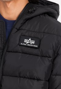 Must musta jakk, millel on nähtav eesmine tõmblukk, kapuuts ja ristkülikukujuline Alpha Industriesi logo patch rinnal.
