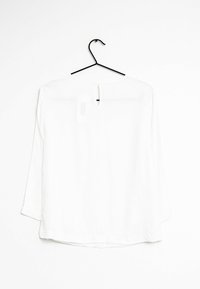 Blusa bianca a maniche lunghe appesa a una gruccia nera contro un muro bianco, vista dal retro con un dettaglio alla scollatura a chiave.