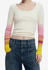 Maglietta a maniche lunghe a costine color crema con maniche a strisce sfumate in rosa, giallo e arancione. Abbinata a jeans in denim blu a vita alta.