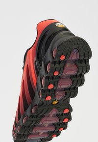 Chaussure de sport rouge et noire avec une tige en maille texturée, dotée d'une semelle en caoutchouc épaisse et d'éléments de rembourrage visibles en rouge et noir.