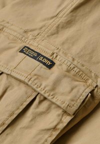 Tessuto color terra con dettagli di cucitura e un'etichetta che mostra "SDRY." La texture sembra liscia e leggermente stropicciata, con una piega visibile.