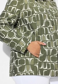 Chaqueta verde con un patrón, hecha de tela con una mezcla de tonos oscuros y claros. Presenta un cierre de botón y un bolsillo lateral.