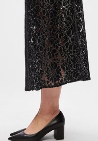 Juoda nėriniuota suknelė su gėlių raštais, pusiau permatoma tekstūra ir tiesia siluetu. Dėvima su juodomis smailuotomis ir storapadėmis bateliais.