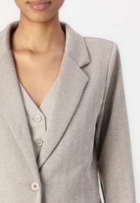 Beige Fischgrätenblazer mit Reverskragen, einer einzelnen Knopfleiste und strukturiertem Stoff, darüber ein hellgraues Oberteil mit Knöpfen.