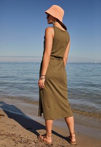 Robe sans manches vert olive avec une ceinture à la taille, mi-longue, en tissu texturé. Associée à un chapeau seau pêche et des sandales à lanières.