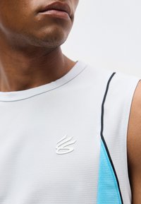 Κοντινό πλάνο ενός ατόμου που φοράει λευκό αθλητικό τοπ χωρίς μανίκια με γαλάζιες και μαύρες λεπτομέρειες και μικρό λευκό λογότυπο στο στήθος.