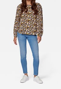Blusa floreale a maniche lunghe con motivi gialli, rosa e bianchi su uno sfondo nero; abbinata a jeans skinny blu e sneakers bianche.