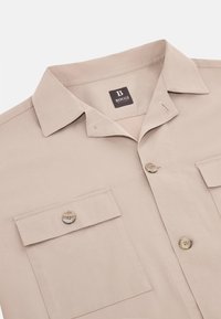Camicetta beige a maniche corte in cotone. Caratterizzata da un colletto, due tasche sul petto con chiusure a bottone e un'etichetta con logo all'interno del colletto.