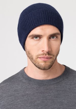 Bonnet en tricot côtelé bleu marine en laine douce, design ajusté, porté par une personne aux cheveux courts, associé à un pull gris.