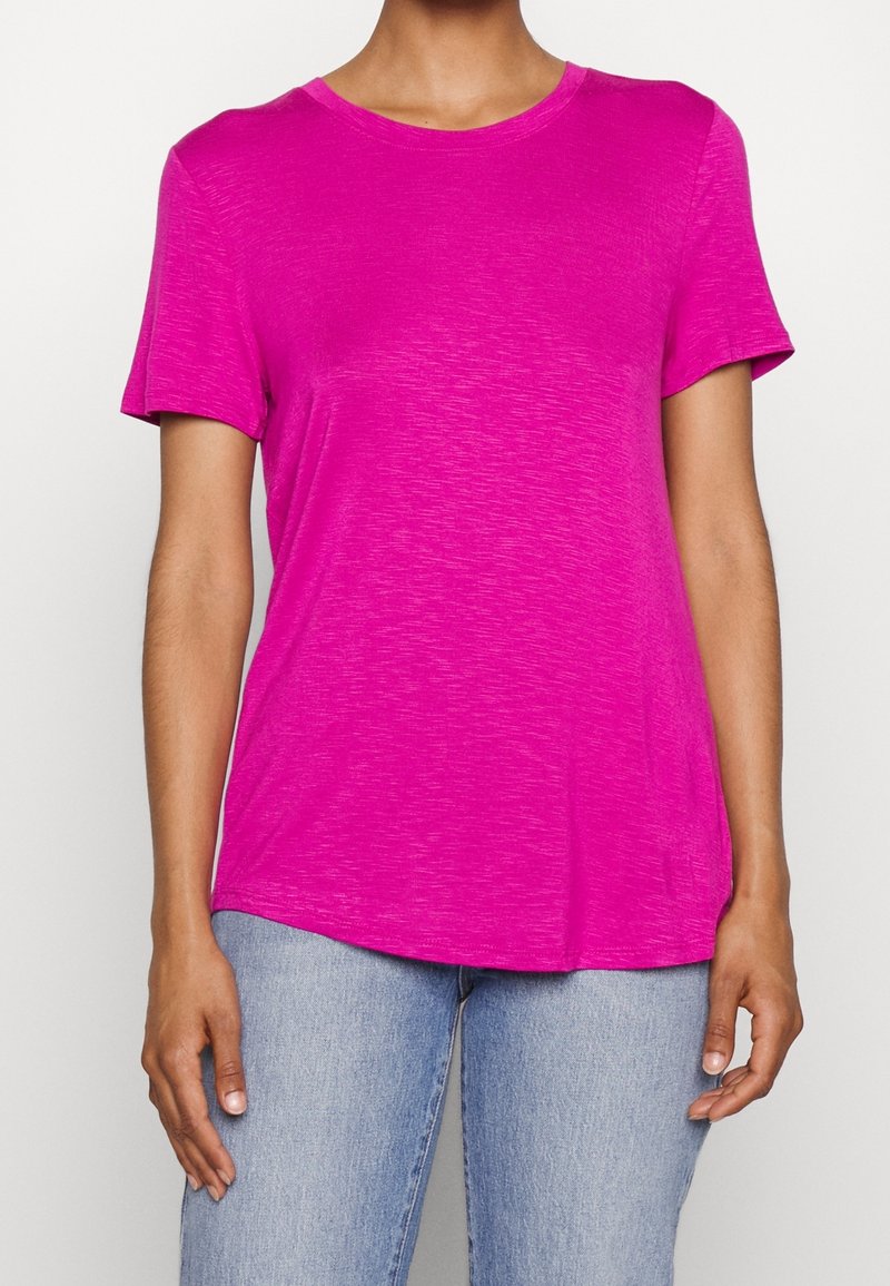 Personne portant un t-shirt rose vif à manches courtes avec col rond et un jean bleu clair, debout devant un fond blanc uni.