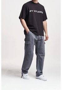 Mann trägt schwarzes "2Y STUDIOS" T-Shirt, graue Cargo-Hosen mit Seitentaschen und weiße Sneakers vor hellem, einfarbigem Hintergrund.