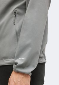 Gros plan sur un bras portant une veste grise avec une poche zippée et une étiquette "WINDWALL" près du poignet.