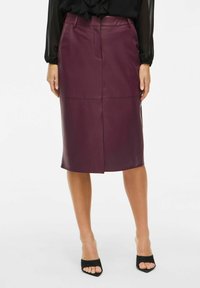 Žena v sukni z plnej kože do kolien v farbe slivky, skombinovanej s čiernou priehľadnou blúzkou a čiernymi sandálmi na odkryté prsty.