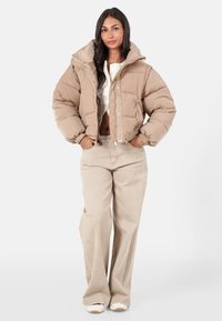 Beige Daunenjacke mit überschnittenem Kragen und elastischen Bündchen, kombiniert mit hellbeigen weit geschnittenen Hosen und weißen Sneakers. Glatte, gestepfte Textur.