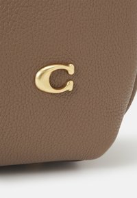 Surface en cuir taupe texturée avec un petit emblème métallique doré en forme de lettre "C" dans le coin inférieur droit.