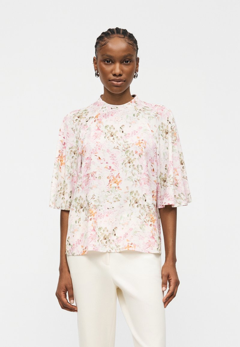 Femme aux cheveux tressés portant un chemisier ample à manches courtes avec un motif floral pastel et un pantalon blanc sur un fond uni.