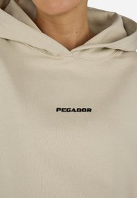 Μπεζ φούτερ από μαλακό ύφασμα με μπροστινή τσέπη, κουκούλα με κορδόνι και λογότυπο "PEGADOR" σε μαύρη κεντητή εκτύπωση στο στήθος.