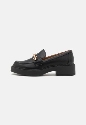Mocassin en cuir noir avec un bout arrondi, des surpiqûres visibles et un ornement chaîne doré. Comprend un talon épais de type bloc noir.
