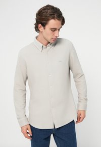 Világos bézs hosszú ujjú ing gombos gallérral, texturált anyagból, elülső zsebbel és halvány logóval a mellkason. Kék farmernadrággal viselve.