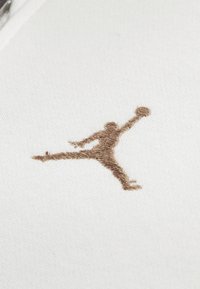Witte stof met een bruin geborduurd Jumpman-logo, waarop een figuur in de lucht met een basketbal in de hand wordt afgebeeld.