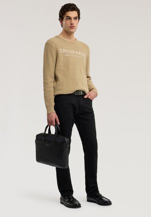 Jeune homme debout, portant un pull beige Trussardi, un pantalon noir, une ceinture noire, des chaussures noires, tenant une mallette noire dans la main gauche.
