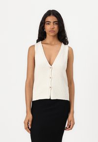 Beżowy dzianinowy kamizelka z dekoltem w serek, z zapięciem na guziki z przodu oraz teksturowanym wykończeniem. Połączona z czarną prążkowaną spódnicą bez rękawów.