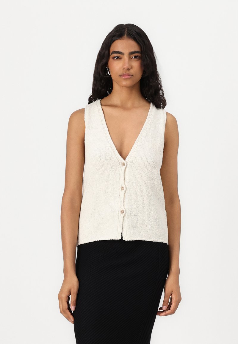 Beżowy dzianinowy kamizelka z dekoltem w serek, z zapięciem na guziki z przodu oraz teksturowanym wykończeniem. Połączona z czarną prążkowaną spódnicą bez rękawów.