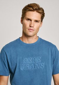 Mėlynas medvilninis trumpomis rankovėmis marškinėliai su iškilniu tekstu "PEPE JEANS" ant priekinės pusės. Atsipalaidavęs siluetas ir lygus tekstūra.