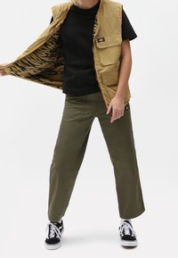 Person med svart t-shirt, beige väst med tigerrandigt foder, olivgröna byxor och svart-vita sneakers står med benen isär.