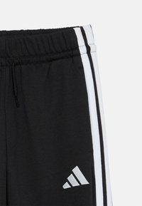 Calças atléticas pretas com riscas brancas nas laterais, apresentando uma cintura elástica e cordão. Logótipo prateado na perna esquerda.