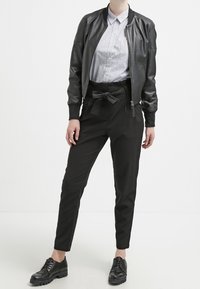 Veste bomber en cuir noir portée sur une chemise rayée à col, associée à un pantalon noir ajusté avec une ceinture nouée, et des chaussures noires à lacets.